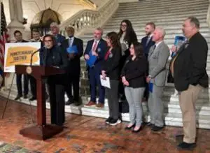 Felicity A. Williams, the executive director of the Pennsylvania Policy Center, speaks at a lectern in the rotunda of the PA state Capitol building. PA legislators and another PPC director, Dwayne Heisler, stand behind her. 